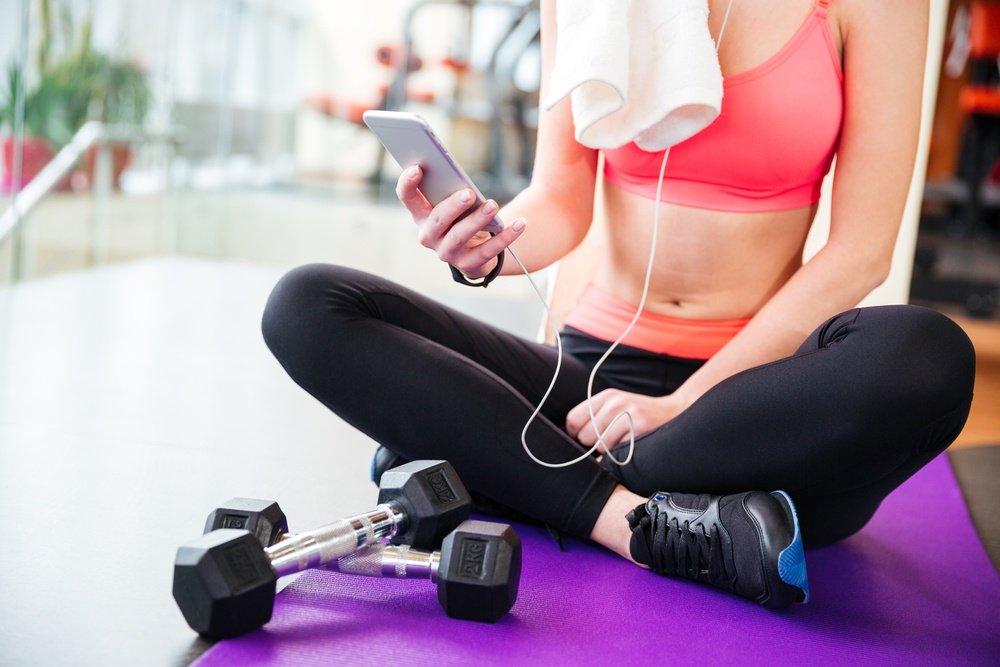 Closeup of young woman athlete sitting on mat and using mobile phone in gym